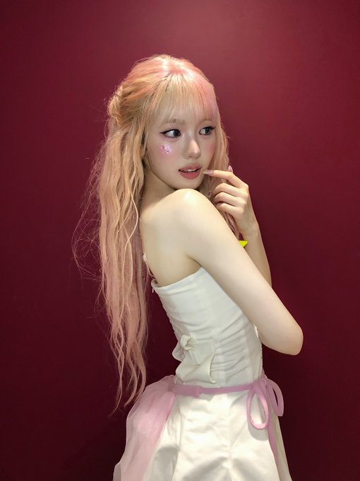 A young woman with long pink/peach hair poses against a dark red background, wearing a white dress with a pink sash, with glittery makeup on her cheeks.