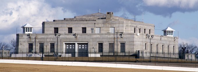 The image features a large, gray concrete building with multiple windows and a prominent tower at its center. The building appears to be an airport or a similar facility, as indicated by the presence of several airplanes visible in the background. The sky above is cloudy, suggesting that the weather might not be ideal for flying.
