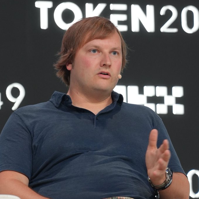 A man with light brown hair speaks into a headset microphone while wearing a dark blue polo shirt, with 'TOKEN 2049' partially visible in the background.