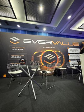 An indoor booth display for "EVERVALUE" features a large illuminated logo, marketing materials, and tables with chairs arranged for visitors.