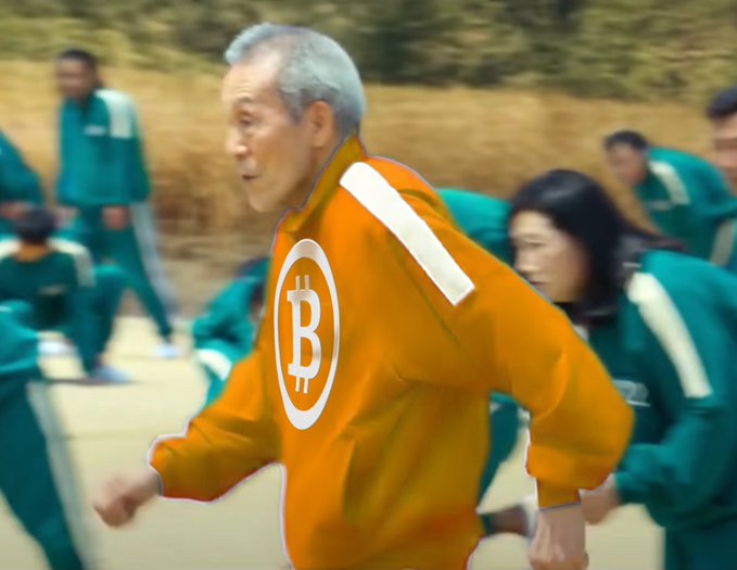 The image features a man wearing an orange jacket with a white "B" logo, running through a field of tall grass. He appears to be in motion as he runs past other people who are also present in the scene. The background includes trees, suggesting that they might be in a rural or natural setting. The post caption reads: "btc on the move". This phrase is likely meant to reference Bitcoin, which is often associated with the term 'bitcoin' due to its association with digital currency and blockchain technology.