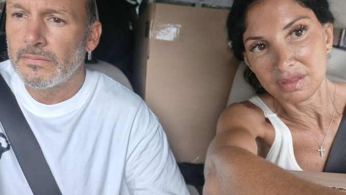 A man and woman sitting in a vehicle with seats covered in cardboard, both wearing seatbelts, looking forward with serious expressions.