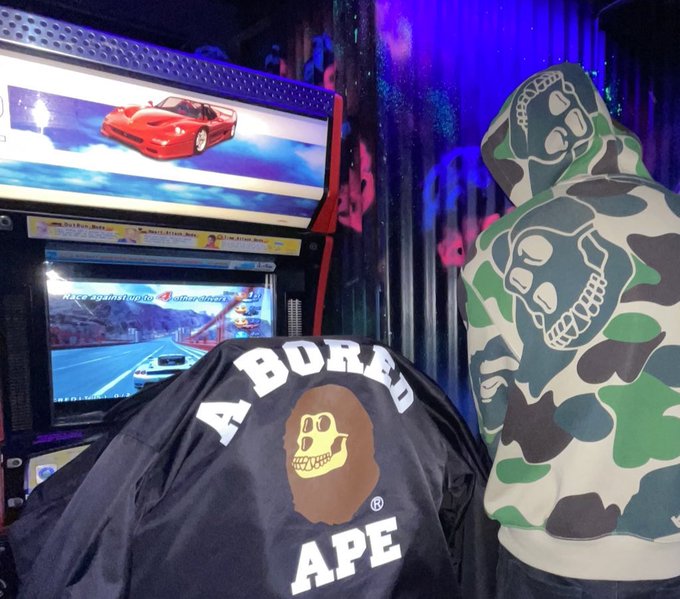 The image features a person wearing a black jacket with a white "A" on it, which has the word "Abored" written on it. The jacket appears to be made of a shiny material that reflects light. In front of this individual is an arcade game featuring a car and a skull design. The background shows a blue wall adorned with graffiti, giving the scene a unique and edgy atmosphere. The post related to the image reads "Bored Ape Yacht Club", suggesting that the person in the jacket might be interested in or associated with the arcade game or the Bored Ape Yacht Club event.