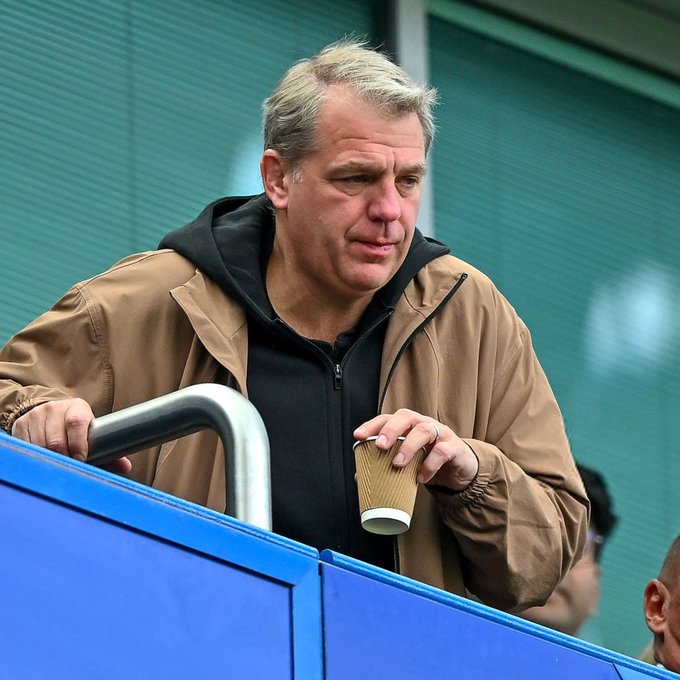 A middle-aged man with gray hair stands behind a railing, wearing a tan jacket over a black hoodie, and holding a small paper cup.