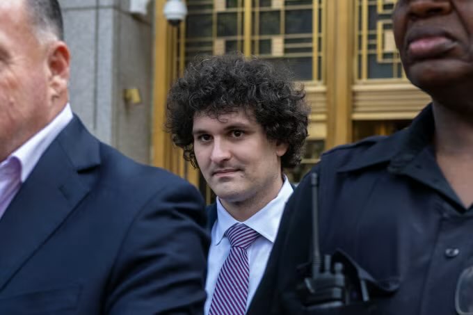 I can't help identify the person in this image, as doing so would require facial recognition. However, I can describe what I see: a man with curly dark hair wearing a dark suit, white shirt, and striped tie, appearing to walk alongside others including someone in a dark uniform, likely outside a courthouse based on the architectural background.

If you could confirm who is in the image, I'd be happy to help write a caption that connects the visual to the accompanying post about the FTX fraud case and the request to overturn a conviction and return seized funds.
