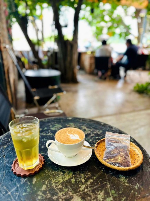A latte art coffee, iced tea, and a small basket of dried tea sit on a dark stone table at an outdoor café. The moody, overcast atmosphere matches the caption's reflection on Hanoi's gloomy weather, offering a quiet moment amid the cloudy day.