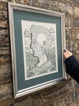 A framed artwork held against a stone wall, depicting a profile silhouette of a head filled with intricate labyrinth and tree patterns.