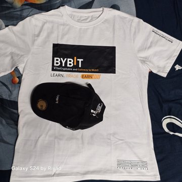 The image features a white t-shirt laid out flat, with a black baseball cap placed next to it. The t-shirt has a logo for Bybit on it, indicating that it's a merchandise item related to cryptocurrency or web3. The cap is positioned in such a way that it partially covers the t-shirt, creating an interesting visual effect. The caption below the image reads "Small gift, big motivation! Today received this awesome Bybit merch package, loving the clean design and crypto vibe!".