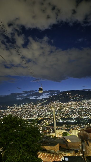 A nighttime view of a sprawling city illuminated with thousands of lights, with a cable car suspended on lines crossing the foreground under a cloudy sky.