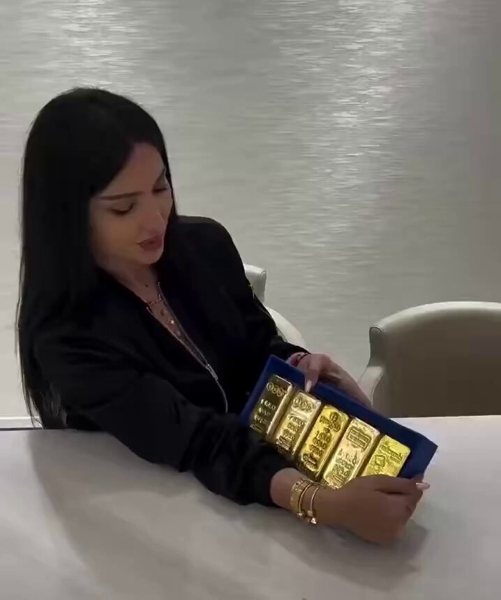 The image features a woman sitting at a table, holding a gold bar with her hands. The gold bar has four distinct pieces of jewelry attached to each end, adding an elegant touch to its appearance. The woman appears to be examining or admiring the gold bar closely.