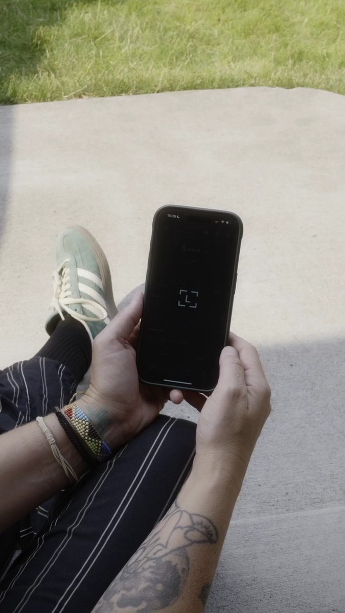 A person with tattooed arms and beaded bracelets holds a smartphone displaying a loading screen, with their green sneakers visible as they sit on a blanket outdoors. The image visually reinforces the post's message by depicting someone using the Ledger Wallet app on their phone to access Somnia assets.