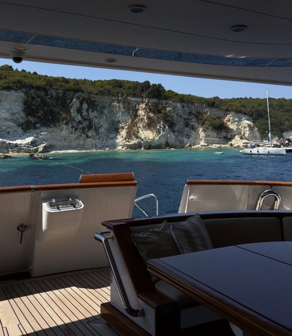 View from the deck of a luxury yacht looking out at turquoise Mediterranean waters and white limestone cliffs with a sailboat anchored nearby.