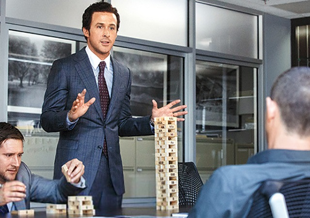 A man in a blue pinstripe suit and tie stands at a conference table gesturing with his hands while speaking, with a tall Jenga-style wooden block tower on the table and colleagues seated around him.