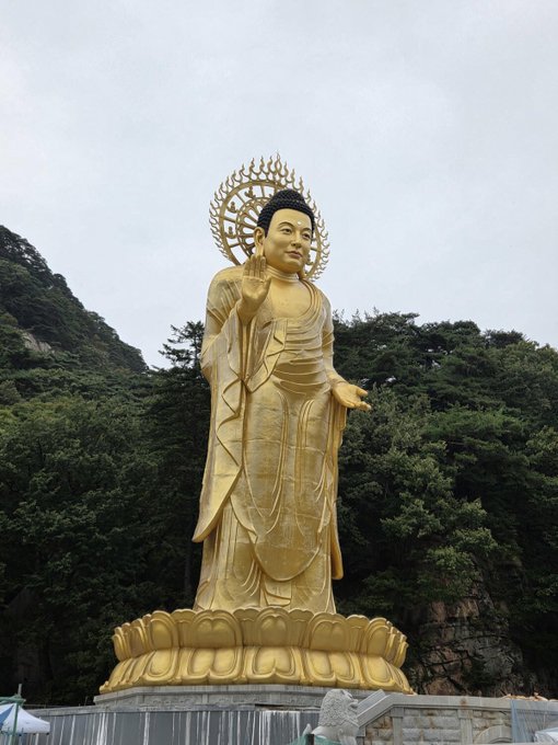 A tall golden Buddhist statue stands on a lotus pedestal in front of a forested mountainside under a cloudy sky.