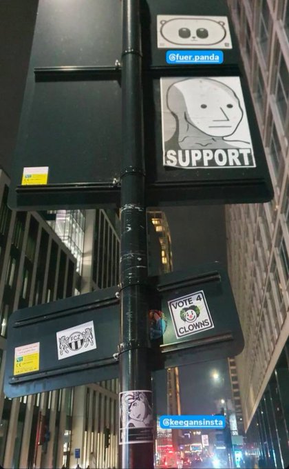 A nighttime urban scene shows a street pole covered in stickers, including ones reading "SUPPORT" with a monk illustration, "VOTE 4 CLOWNS," and the handles "@fuer.panda" and "@keegansinsta." The "$NPC" hashtag in the post references the meme, with the stickers' absurd, generic humor—particularly the "Vote 4 Clowns" tag—embodying the non-player character vibe of unthinking, meme-driven internet culture plastered across the city.
