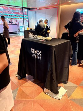A black "OKX Wallet" promotional table is set up in a bustling event space, with branding clearly displayed on the tablecloth as people gather around in the background. The "Enjoy the moment" caption captures the lively, social atmosphere of this crypto networking event.