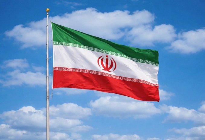 The flag of Iran waves against a blue sky with white clouds, displaying its green, white, and red horizontal stripes with the central emblem.