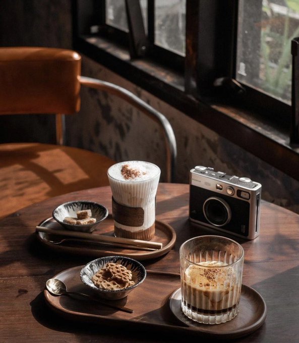 A cozy scene on a wooden table featuring a layered coffee drink, a glass of coffee with cream, small bowls of cookies and treats, and a vintage-style camera, all illuminated by sunlight from a nearby window.