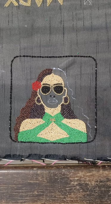 A rhinestone art piece depicting a woman with sunglasses, hoop earrings, a red flower in her hair, and a green halter top, displayed on a dark fabric background.