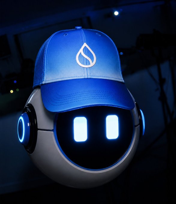 A spherical robot mascot with glowing blue rectangular eyes wears a blue cap featuring the Sui water-droplet logo, visually representing the Suiball hardware wallet highlighted in the accompanying post about its security components.
