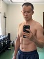 A shirtless young man in blue shorts stands in what appears to be a gym, holding a smartphone to take a mirror selfie.