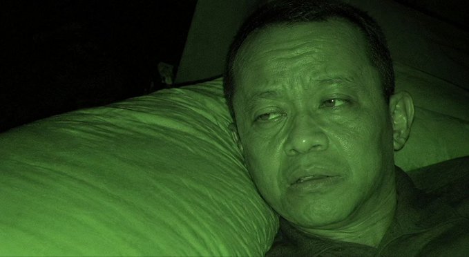 A man lies in bed with a serious expression, captured in a green-tinted low-light photo against a striped pillow.