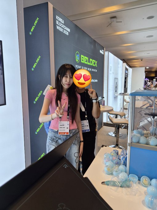 Two young women posing for a photo at the 'KOREA BLOCKCHAIN WEEK 2025' event in front of a 'BITLDEX' banner, with one face obscured by a heart-eyes emoji, and display cases containing blue capsules visible on the right.