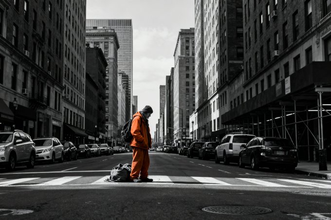 A black-and-white street photograph of a person in a bright orange jacket and backpack standing on a crosswalk in the middle of an empty city street lined with tall buildings and parked cars.