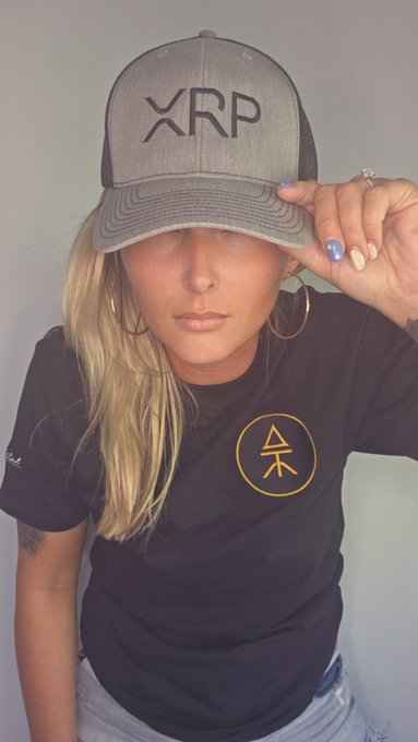 A selfie-style photo of a woman wearing a gray 'XRP' branded cap and black t-shirt featuring an orange crypto-style logo, against a plain light-colored background.