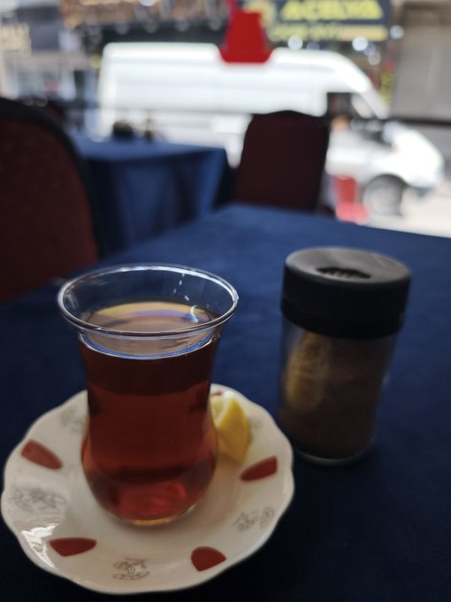 A tulip-shaped glass of dark tea sits on a decorative saucer beside a metal sugar shaker on a navy-blue tablecloth, with a blurred teahouse interior in the background—setting the scene for the newly opened AKD Teahouse mentioned in the post.