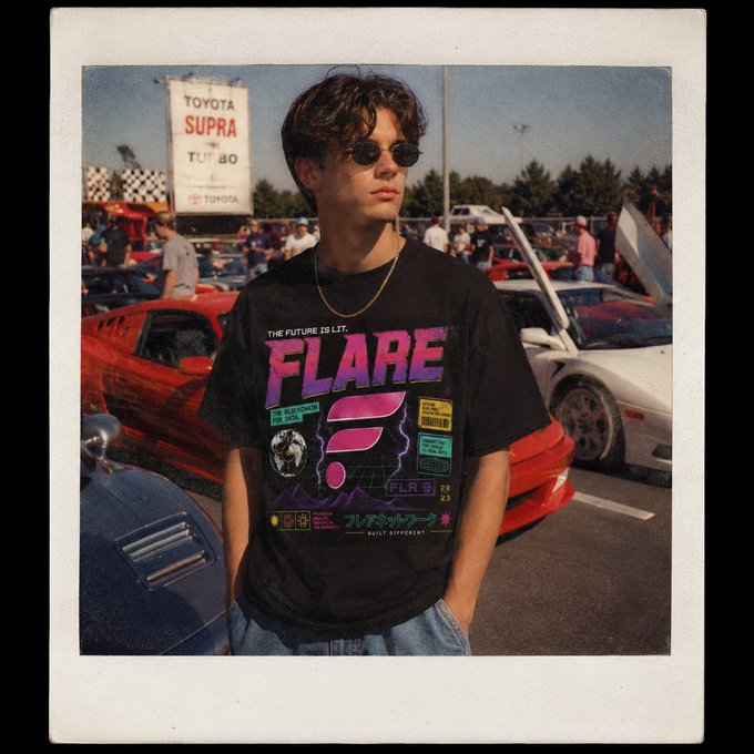 The image features a young man wearing a black t-shirt with "Flare" written across the front, standing next to a red car. The background of the photo shows several other cars parked nearby, including a white car that appears to be a Toyota Supra. There are also people present in the scene, some of whom might be attending an event or gathering related to the cars on display.