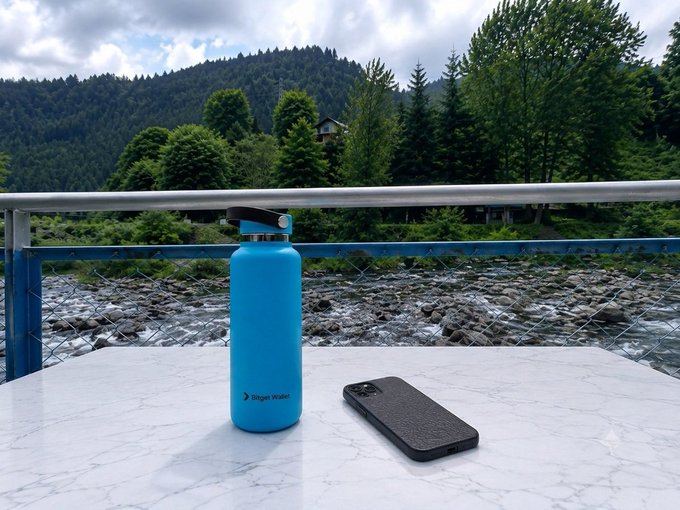 A blue "Bigger Walker" water bottle and a smartphone rest on a marble table overlooking a tranquil river and forested mountains, perfectly capturing the balance of nature and connectivity in weekend mode.
