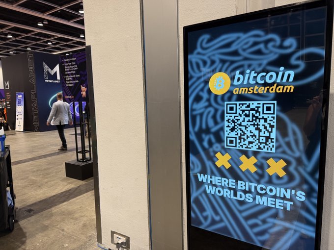 A digital display at an event advertises "bitcoin amsterdam" with the tagline "WHERE BITCOIN'S WORLDS MEET" and features a QR code and Amsterdam's XXX symbol.