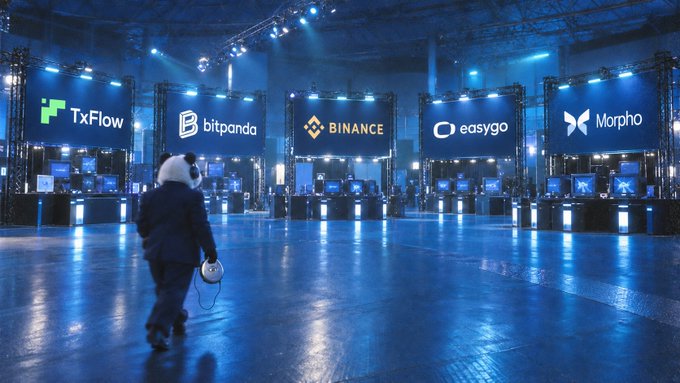 A bear mascot in a suit walks through a blue-lit crypto conference venue with illuminated sponsor signs including 'TxFlow', 'bitpanda', 'BINANCE', 'easygo', and 'Morpho'.