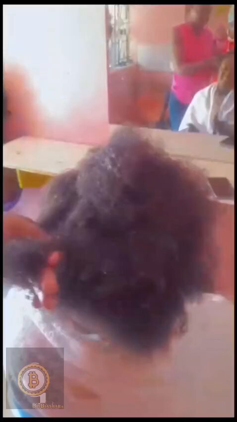 The image shows a person with long, dark hair sitting in front of a mirror at a salon. The person's hair appears to be wet or recently washed, as there are water droplets visible on their head. In the background, another person is also present, possibly attending to the client or waiting for their turn.