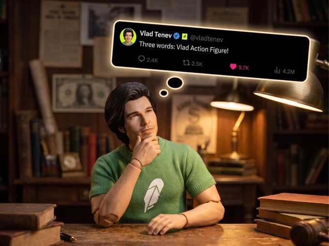 A young man in a green sweater sits thoughtfully at a wooden desk in a study, thinking about 'Three words: Vlad Action Figure!' as shown in a tweet from Vlad Tenev (@vladtenev) with 2.4K replies and 9.7K likes.