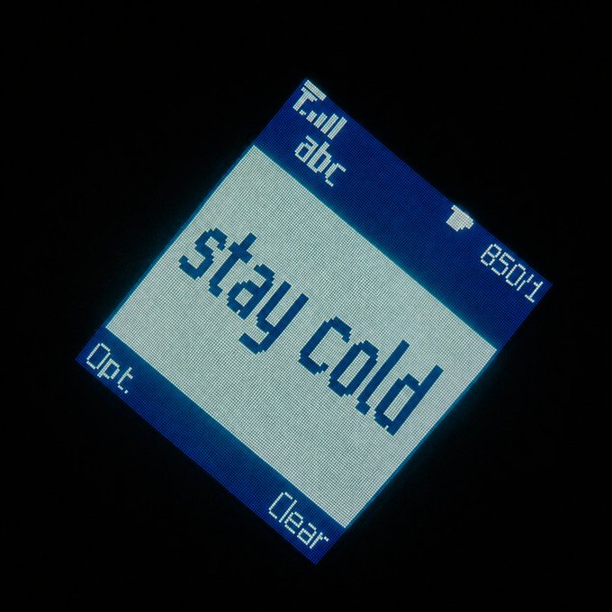 A retro-style mobile phone screen displays the message "Stay cold" in pixelated blue text, with options to "Dial," "abc," "Opt.," and "Clear," alongside a battery icon and the time "8:50." The image reinforces the post's message about secure cold wallet storage, with "Stay cold" serving as a nod to both literal cold storage and the warning to keep your offline crypto assets safely hidden and backed up.