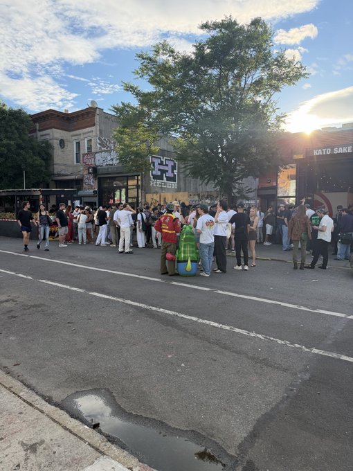 The image shows a group of people gathered outside, possibly attending an event or gathering. There are several individuals standing around, some closer to the foreground while others are further back. A few people can be seen carrying handbags as they mingle with one another. The scene is set against a backdrop of buildings and trees, creating an urban atmosphere.

The post on social media indicates that this event might be related to a block party or similar gathering taking place at the Eth Bushwick block in New York City. The image captures the essence of community engagement and social interaction within an urban setting.