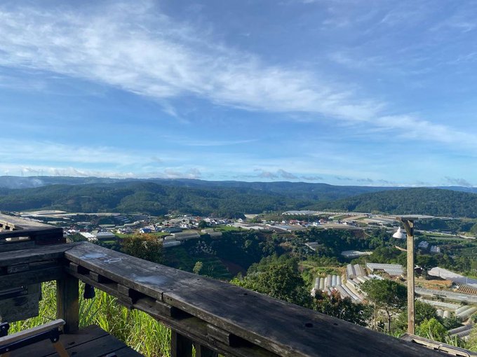 A scenic landscape photograph taken from an elevated viewpoint showing green rolling hills, agricultural fields with white greenhouses, distant mountains, and a blue sky with scattered clouds, with a wooden railing in the foreground.