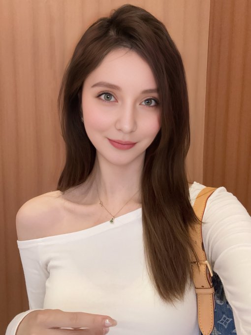 A woman with long brown hair and green eyes smiles for a portrait, wearing a white off-the-shoulder top and a delicate pendant necklace.