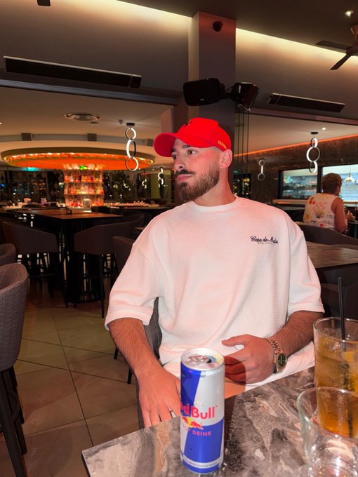 A man wearing a red cap and a "Côte d'Azur" sweatshirt sits at a marble table in a dimly lit bar with a Red Bull Drink can and iced beverages in front of him, looking off to the side. The post promises a 2 SOL prize to the best caption within 3 hours.