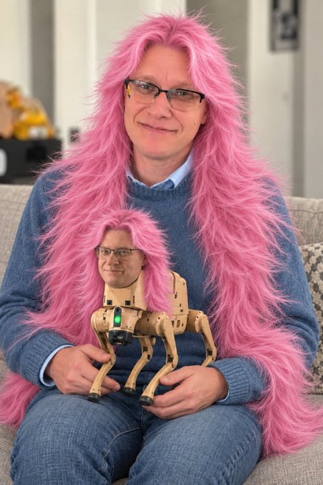 A person wearing a long pink wig and glasses holds a small robotic quadruped that has a matching pink wig and a printed face, creating a humorous duplicate effect.