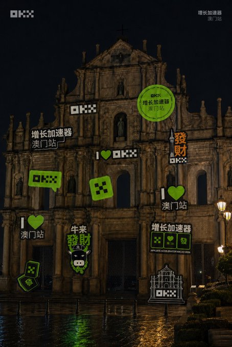 The Ruins of Saint Paul's in Macau is illuminated at night, decorated with green "OKX" cryptocurrency brand stickers and Chinese text reading "增長加速器 澳門站" (Growth Accelerator Macau Station). The image ties to the "GM💚 May the winds of wealth blow your way!" post by promoting OKX's Macau crypto event with cheerful green branding and wealth-themed messaging.