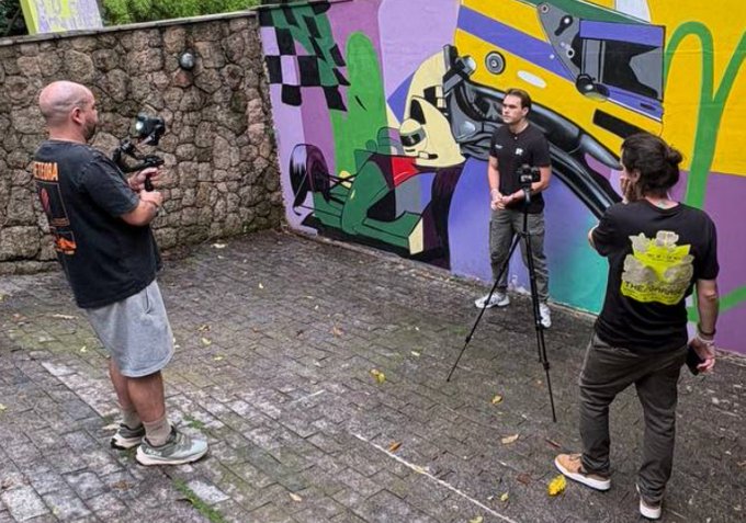 Three men film a video in a cobblestone area in front of a colorful racing-themed mural, with one operating a camera on a tripod and another wearing a shirt with "The Grumps" logo.