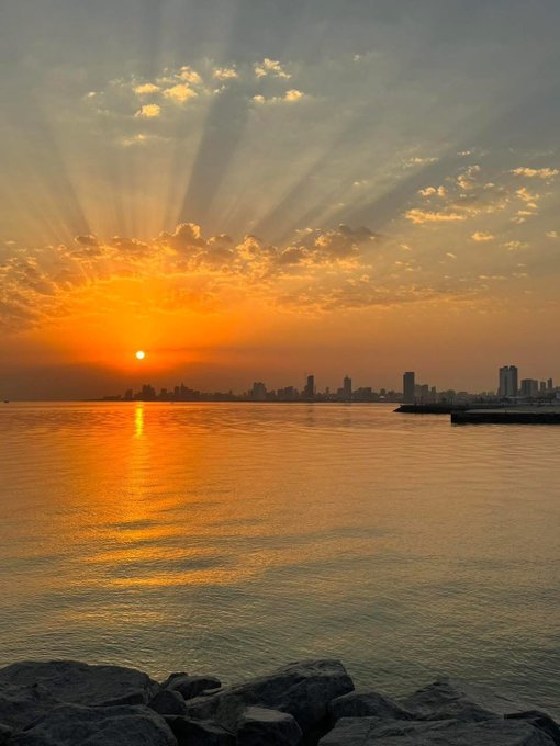 Photograph of a vibrant sunset over a city skyline across water, with golden sun rays radiating through scattered clouds and the sun reflected on the calm sea surface.