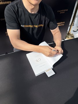 A person wearing a black t-shirt with 'EXCHANGE THE WORLD' and 'CHANGEPHAO' printed on it signs a book at a table.