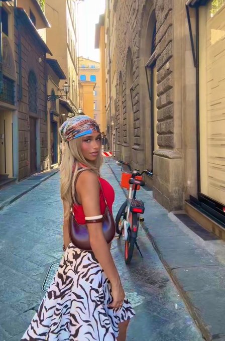 A blonde woman wearing a colorful headscarf, red top, and zebra-print skirt stands in a narrow European-style cobblestone street with an orange rental bicycle behind her.