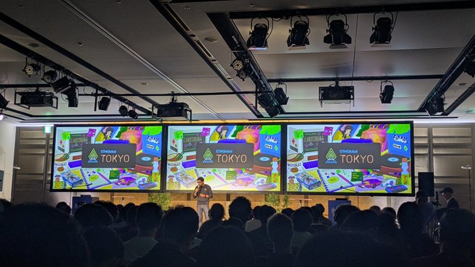 A conference audience watches a speaker on stage with three large screens displaying '2025 AI STRATEGIES IN TOKYO' above the presentation.