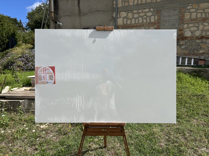 A blank white canvas or board wrapped in plastic sits on a wooden easel in a grassy area, with a stone building, trees, and a small product label visible in the background.