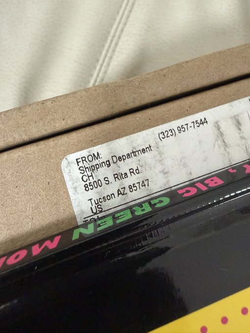 A shipping label on a cardboard box shows a return address from "Shipping Department" at 8500 S. Rita Rd., Tucson AZ 85747, with phone number (323) 957-7544, resting against a black box with colorful text that appears to read "BIG GREEN" in pink and green lettering. The label's Tucson, Arizona address and partial branding visible suggest a Chrome Hearts package arrival, though the specific return sender details don't directly reveal the luxury brand.